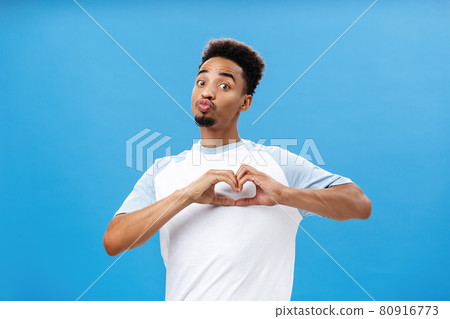 Funny and cute dark-skinned young male student in t-shirt folding lips in mwah or kiss showing heart gesture over breast giving love and affection signs to girl he likes over blue wall Funny and cute dark-skinned young male student in t-shirt folding lips in mwah or kiss showing heart gesture over breast giving love and affection signs to girl he likes over blue wall 80916773