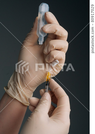 Vertical close-up studio shot of unrecognizable doctor in white gloves holding and filling up vaccine to syringe on black isolated background. 80917029
