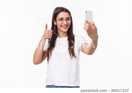 Communication, technology and lifestyle concept. Portrait of upbeat happy, smiling caucasian girl in glasses, internet blogger taking selfie, record video message to followers thumbs-up, hold phone 80917107