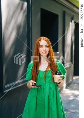 Vertical medium shot portrait of attractive young woman in green dress holding takeaway coffee cup, using mobile phone walking at city street 80917740