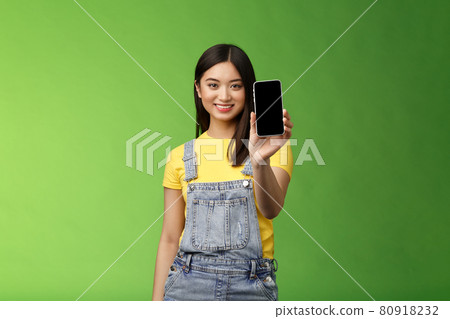Confident assertive good-looking female asian introduce smartphone app, extend hand hold telephone arm, smiling delighted, recommend social profile, bragging own blog, green background 80918232
