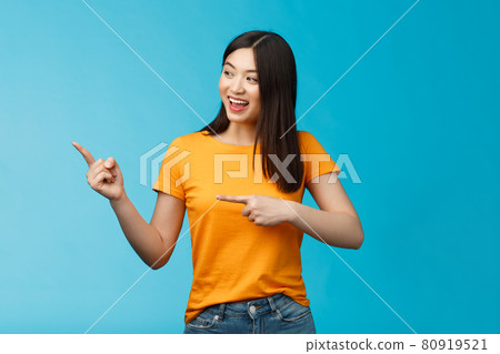 Friendly enthusiastic asian girl curiously looking sideways, pointing left interesting object, grinning joyfully discuss new opened store, stand blue background carefree lively talking 80919521
