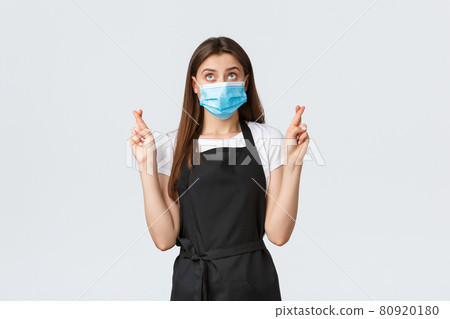 Covid-19 social distancing, cafe employees, coffee shops and coronavirus concept. Hopeful cute waitress, cashier or saleswoman in black apron and medical mask making wish with fingers crossed Covid-19 social distancing, cafe employees, coffee shops and coronavirus concept. Hopeful cute waitress, cashier or saleswoman in black apron and medical mask making wish with fingers crossed 80920180