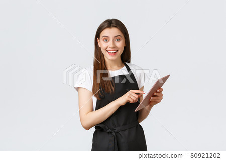 Grocery store employees, small business and coffee shops concept. Friendly cute female barista in black apron smiling at camera, using digital tablet to manage cafe orders Grocery store employees, small business and coffee shops concept. Friendly cute female barista in black apron smiling at camera, using digital tablet to manage cafe orders 80921202