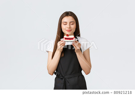 Grocery store employees, small business and coffee shops concept. Female cafe worker in black apron holding tasty cake and smelling delicious dessert with pleased smile and closed eyes 80921218