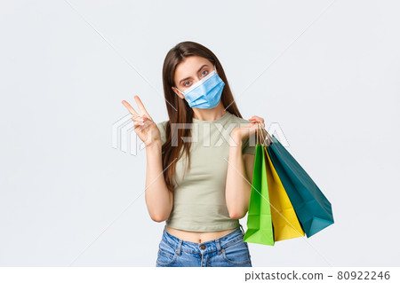 Social distancing, coronavirus and shopping, fashion concept. Silly cute stylish woman in medical mask shopping in malls during covid-19, show peace sign and bags with purchased items Social distancing, coronavirus and shopping, fashion concept. Silly cute stylish woman in medical mask shopping in malls during covid-19, show peace sign and bags with purchased items 80922246