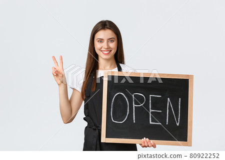 Grocery store employees, small business and coffee shops concept. Friendly cafe worker in black apron show peace gesture and we are open sign, smiling as inviting people visit shop or cafe Grocery store employees, small business and coffee shops concept. Friendly cafe worker in black apron show peace gesture and we are open sign, smiling as inviting people visit shop or cafe 80922252