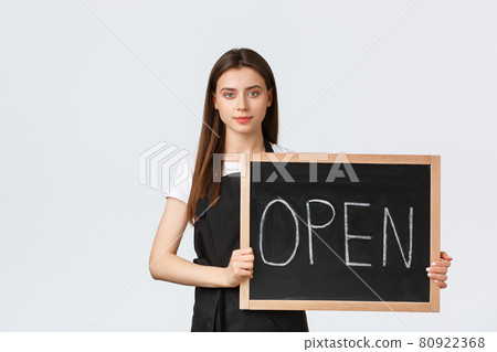 Grocery store employees, small business and coffee shops concept. Cute young female shop worker in black apron holding we are open sign, inform guests cafe is working, white background Grocery store employees, small business and coffee shops concept. Cute young female shop worker in black apron holding we are open sign, inform guests cafe is working, white background 80922368