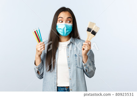Social distancing lifestyle, covid-19 pandemic, self-isolation hobbies and leisure concept. Excited creative asian girl in medical mask showing coloured pencils and painting brushes, want draw Social distancing lifestyle, covid-19 pandemic, self-isolation hobbies and leisure concept. Excited creative asian girl in medical mask showing coloured pencils and painting brushes, want draw 80923531