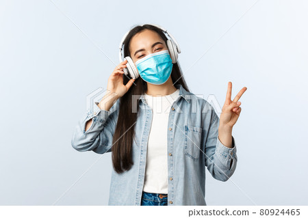 Social distancing lifestyle, covid-19 pandemic and self-isolation leisure concept. Happy smiling asian girl listening music in wireless headphones, wear medical mask and show peace sign Social distancing lifestyle, covid-19 pandemic and self-isolation leisure concept. Happy smiling asian girl listening music in wireless headphones, wear medical mask and show peace sign 80924465
