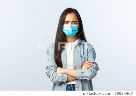 Social distancing lifestyle, covid-19 pandemic life and leisure concept. Suspicious and disappointed young girl in medical mask, asian female looking displeased, cross arms chest and frowning Social distancing lifestyle, covid-19 pandemic life and leisure concept. Suspicious and disappointed young girl in medical mask, asian female looking displeased, cross arms chest and frowning 80924467