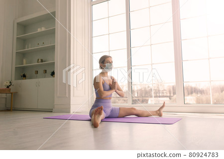 Attractive female sport fitness coach wearing white medical mask do exercises on yoga mat at home. Attractive female sport fitness coach wearing white medical mask do exercises on yoga mat at home. 80924783