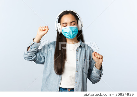 Social distancing lifestyle, covid-19 pandemic and self-isolation leisure concept. Happy upbeat asian girl in medical mask, listening music in wireless headphones and dancing 80924959