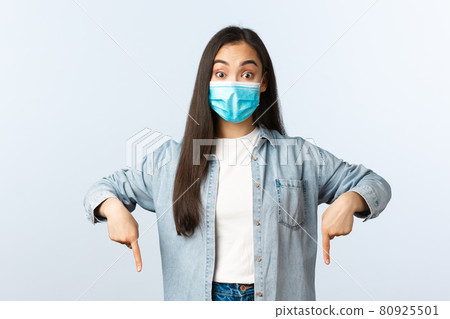 Social distancing lifestyle, covid-19 pandemic everyday life and leisure concept. Surprised happy asian girl found something cool, pointing fingers down to show promo, wear medical mask Social distancing lifestyle, covid-19 pandemic everyday life and leisure concept. Surprised happy asian girl found something cool, pointing fingers down to show promo, wear medical mask 80925501