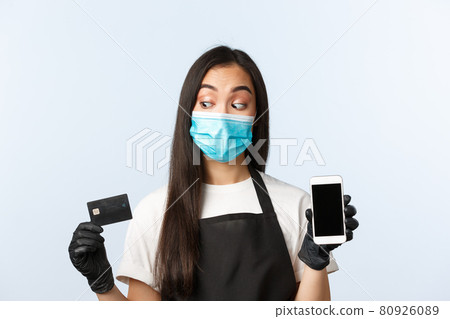 Covid-19 pandemic, coffee shop, small business and preventing virus concept. Intrigued cute asian female cafe owner, barista looking at credit card and holding mobile phone Covid-19 pandemic, coffee shop, small business and preventing virus concept. Intrigued cute asian female cafe owner, barista looking at credit card and holding mobile phone 80926089