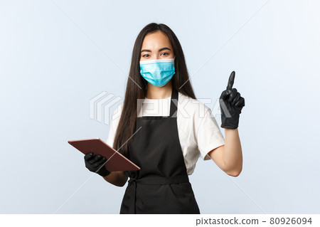 Covid-19, online orders, small coffee shop and preventing virus concept. Smiling friendly asian waitress, female barista in medical mask and gloves point upper right corner, use digital tablet Covid-19, online orders, small coffee shop and preventing virus concept. Smiling friendly asian waitress, female barista in medical mask and gloves point upper right corner, use digital tablet 80926094