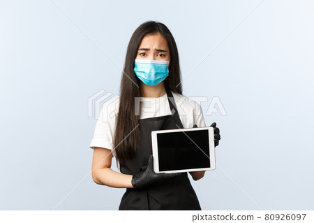 Covid-19 pandemic, social distancing, small business and preventing virus concept. Upset gloomy asian waitress, cafe staff in medical mask and gloves frowning sad and showing digital tablet display 80926097