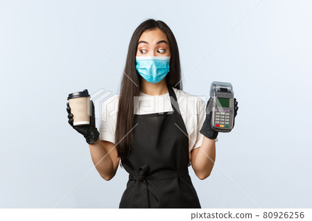 Coffee shop, coronavirus, social distancing and contactless payment concept. Excited asian female barista in medical mask and gloves staring at takeaway cup, holding POS paying terminal Coffee shop, coronavirus, social distancing and contactless payment concept. Excited asian female barista in medical mask and gloves staring at takeaway cup, holding POS paying terminal 80926256