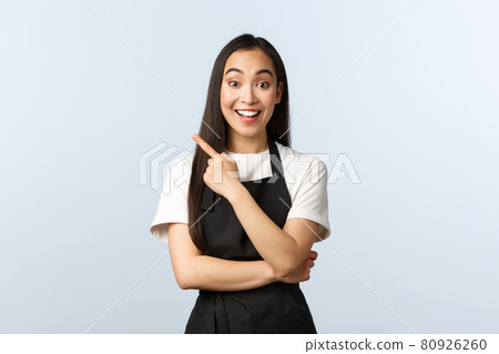 Coffee shop, small business and startup concept. Excited smiling asian female barista, cafe owner in black apron inviting check out new special offers, pointing finger upper left corner at banner 80926260