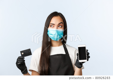 Covid-19 pandemic, coffee shop, small business and virus concept. Thoughtful young asian female barista, waitress in medical mask thinking, holding credit card and show mobile phone display Covid-19 pandemic, coffee shop, small business and virus concept. Thoughtful young asian female barista, waitress in medical mask thinking, holding credit card and show mobile phone display 80926283