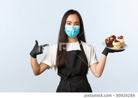 Covid-19, social distancing, small coffee shop business and preventing virus concept. Smiling cute asian female barista, waitress at cafe pointing at muffins desserts, serving order in medical mask Covid-19, social distancing, small coffee shop business and preventing virus concept. Smiling cute asian female barista, waitress at cafe pointing at muffins desserts, serving order in medical mask 80926289
