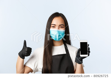 Covid-19 pandemic, coffee shop, small business and preventing virus concept. Smiling attractive asian cafe owner, barista showing smartphone screen and thumb-up, wear medical mask Covid-19 pandemic, coffee shop, small business and preventing virus concept. Smiling attractive asian cafe owner, barista showing smartphone screen and thumb-up, wear medical mask 80926725