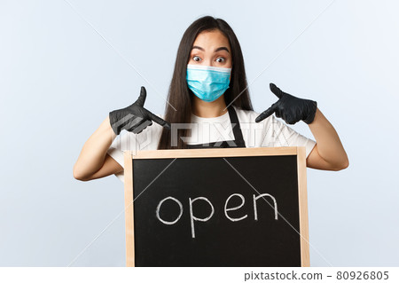 Small business, covid-19 pandemic, preventing virus and employees concept. Excited cute asian female barista, cafe or store worker in medical mask pointing at we are open sign 80926805
