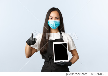 Covid-19, social distancing, small coffee shop and preventing virus concept. Satisfied smiling asian female barista, cafe owner or waitress show thumb-up and digital tablet, wear medical mask 80926839