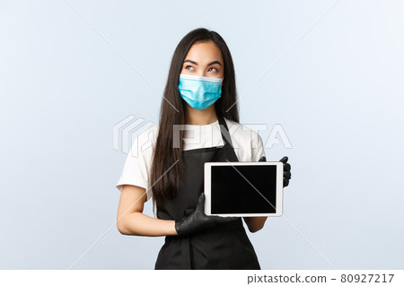 Covid-19, social distancing, small coffee shop business and preventing virus concept. Thoughtful, pleased asian female barista, cafe owner in medical mask and gloves show digital tablet screen Covid-19, social distancing, small coffee shop business and preventing virus concept. Thoughtful, pleased asian female barista, cafe owner in medical mask and gloves show digital tablet screen 80927217