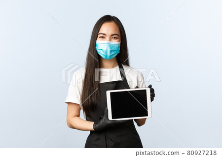 Covid-19 pandemic, social distancing, small business and preventing virus concept. Friendly smiling female asian barista in medical mask and gloves, showing digital tablet, how to make order online Covid-19 pandemic, social distancing, small business and preventing virus concept. Friendly smiling female asian barista in medical mask and gloves, showing digital tablet, how to make order online 80927218