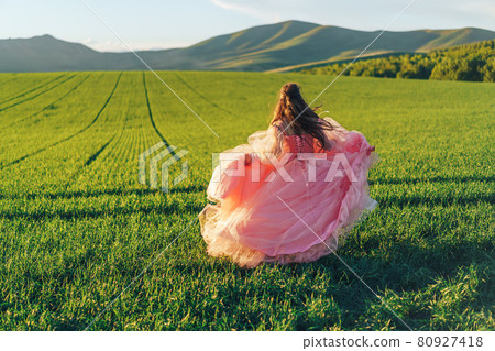Girl in pink dress walking through green fields at sunset 80927418