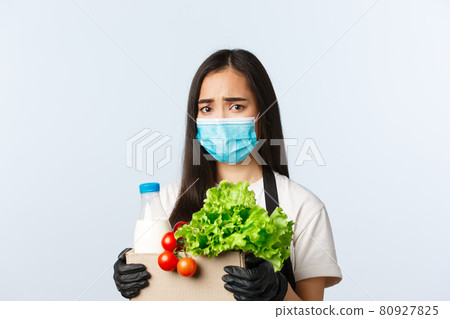 Covid-19, grocery store, employment, small business and preventing virus concept. Worried and upset frowning asian female shop employee, cashier in medical mask holding groceries bag Covid-19, grocery store, employment, small business and preventing virus concept. Worried and upset frowning asian female shop employee, cashier in medical mask holding groceries bag 80927825