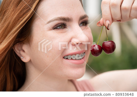 Beautiful young red-haired woman with braces on her teeth eats sweet cherries in the summer outdoors Beautiful young red-haired woman with braces on her teeth eats sweet cherries in the summer outdoors 80933648