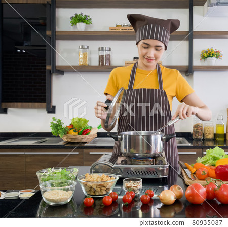 Young asian housewife dressed in an apron and a hair cap, preparing the meat stew in a modern kitchen. Young asian housewife dressed in an apron and a hair cap, preparing the meat stew in a modern kitchen. 80935087