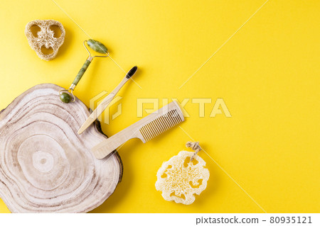 Eco beauty concept. Jade face roller, loofah sponges, toothbrush, wooden comb, wood slice on yellow surface. Set of items for beauty from ecological material. Copy space, flat lay 80935121