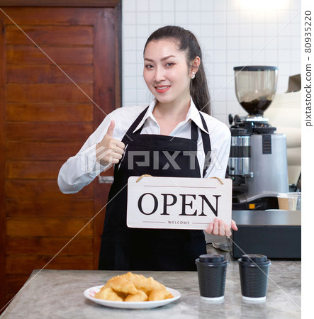 Young asian shopkeeper with a smile holds an OPEN sign in front of a kitchen counter. Morning atmosphere in a coffee shop. 80935220
