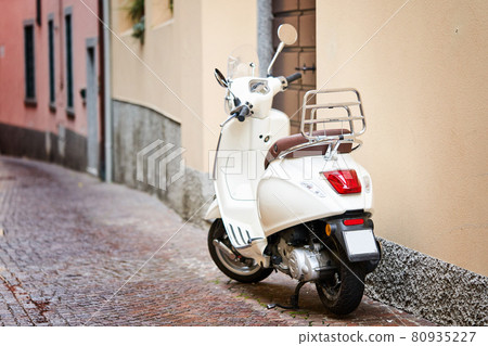 White retro scooter or moped parked on sidewalk of empty narrow street. Motorcycle or motorbike on italian street 80935227