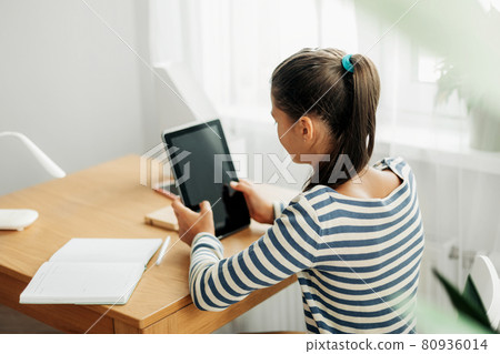 A dark-haired schoolgirl sits at a desk doing her homework using her tablet. Modern technologies, online education, training 80936014