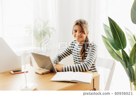 Smiling schoolgirl in a striped sweater sits at the table, does homework on the tablet and takes notes in a notebook. Modern technologies, online education, training 80936021