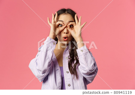 Close-up portrait of amused, speechless and impressed asian young girl seeing something curious and amazing, make glasses with fingers over eyes, fold lips say wow astonished, pink background 80941119