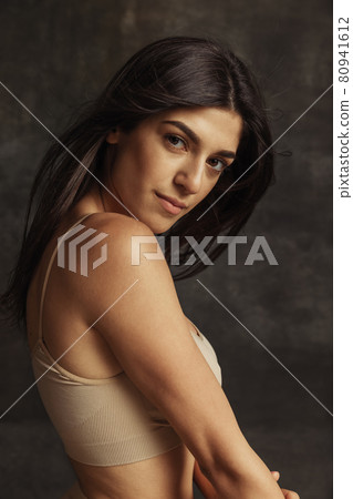 Portrait of young beautiful tanned woman in lingerie posing isolated over dark vintage studio background. Natural beauty concept. 80941612
