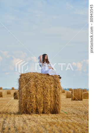 Dreamy young woman sits on the top of the ricks on the field 80943545
