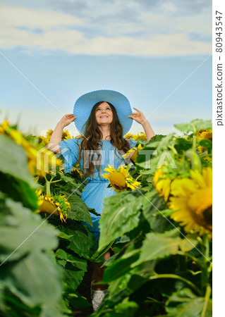 Woman in blue dress holds the edges of straw hat and looking up among sunflowers Woman in blue dress holds the edges of straw hat and looking up among sunflowers 80943547