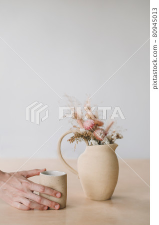 Minimal home decor with dry flowers in a ceramic vase and a cup in female hands. Minimal home decor with dry flowers in a ceramic vase and a cup in female hands. 80945013