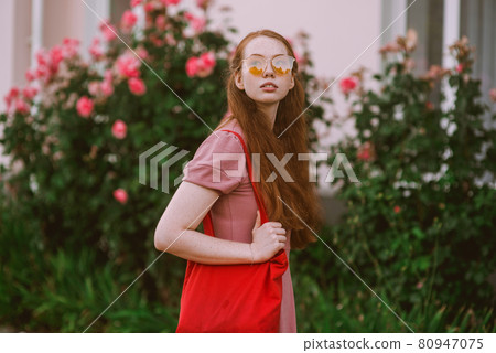Young beautiful red-haired long-haired woman in a summer pink dress walks through the streets of a summer city. Young beautiful red-haired long-haired woman in a summer pink dress walks through the streets of a summer city. 80947075