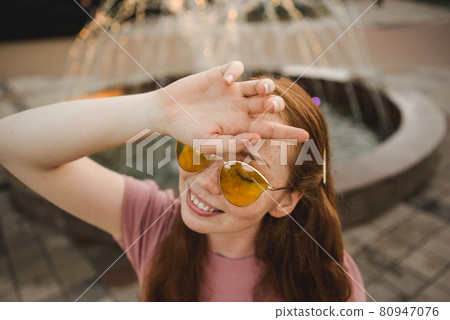 Young beautiful red-haired long-haired woman in a summer pink dress walks through the streets of a summer city. Young beautiful red-haired long-haired woman in a summer pink dress walks through the streets of a summer city. 80947076