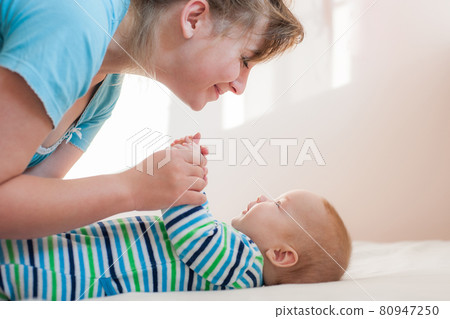 Happy laughing mom and daughter lie on their backs. Happy smiling mother and baby lying on bed at home. Side view Happy laughing mom and daughter lie on their backs. Happy smiling mother and baby lying on bed at home. Side view 80947250