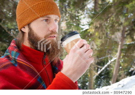 Bearded caucasian man in a hat wrapped in a blanket with a mug of hot drink in a paper cup in a winter coniferous forest on a sunny day Bearded caucasian man in a hat wrapped in a blanket with a mug of hot drink in a paper cup in a winter coniferous forest on a sunny day 80947290