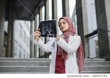 Muslim doctor in hijab, standing outdoor of modern hospital and looking at x-ray image 80947345