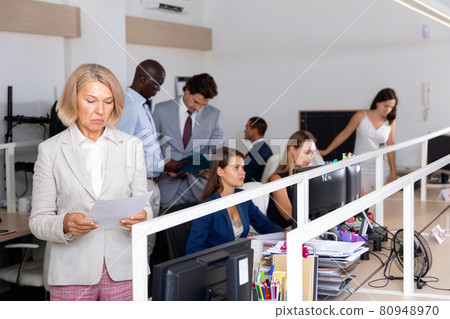 Puzzled and confused mature businesswoman standing with papers in hands in coworking space, thinking about problem to solve 80948970